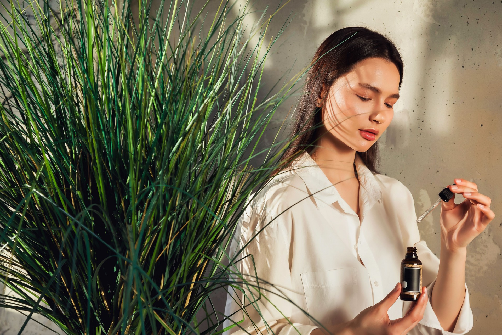 Pretty young lady holding massage oil bottle with pipette in foliage shadow, pensive looking. Lovely woman dripping cosmetic oil at textured wall. Cosmetology and self care concept. Copy ad text space